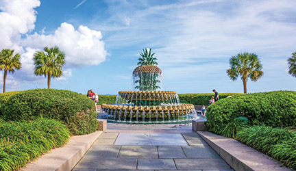 A large pineapple shaped fountain set back along a park walkway with green shrubs on both sides, palm trees in the background under a blue sky with a few white clouds.