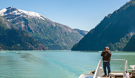A person stands on the deck of a boat, looking across a turquoise lake toward snow-capped mountains under a clear blue sky.