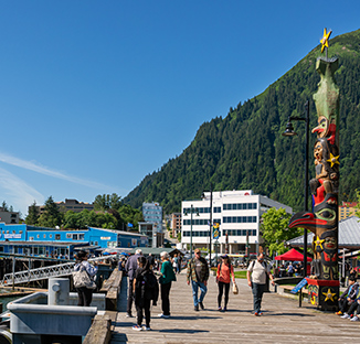 Juneau's boardwalk with people walking, totem pole on the side with buildings and lush, steep mountain just beyond.