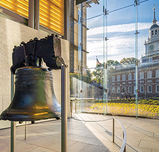 An interior view of the Liberty Bell on its stand inside a modern glass-walled pavilion, with a clear view of Independence Hall and a sunny outdoor courtyard outside.