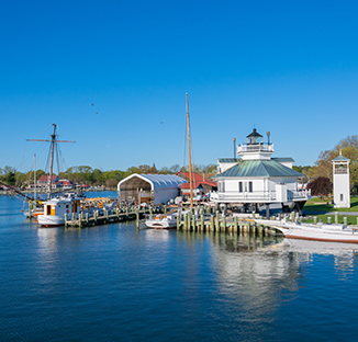 A wide, sunlit view of a calm harbor with several docked boats and traditional sailing vessels next to a distinctive white lighthouse on stilts and boat-building facilities under a clear blue sky.