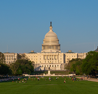 A wide-angle, outdoor shot of the United States Capitol building with its grand neoclassical dome, situated behind a sprawling green lawn with people scattered across the grass under a clear blue sky.