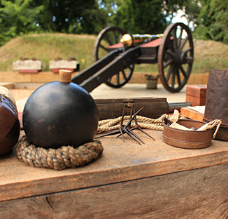 A close-up view of a black round spherical shell resting on a coil of rope on a wooden table, alongside historical artifacts including caltrops, boxes, and a vintage cannon positioned in the background outdoors.