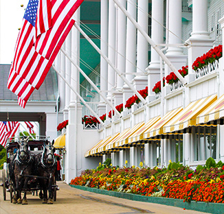 A grand building in Mackinac Island with American flags, horse-drawn carriage, and vibrant flowers.