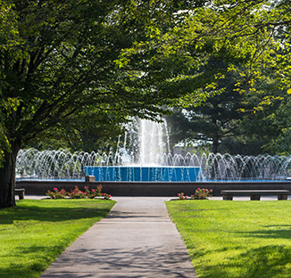 Pathway leading to a large fountain surrounded by trees and benches, in Sault Ste. Marie, MI, with sunlight filtering through the leaves.