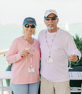 Couple on a small cruise ship deck, wearing casual clothes and lanyards, holding drinks, with ocean and sailboat in background.