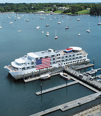 Small cruise ship docked in a harbor with an American flag, surrounded by sailboats and lush greenery.
