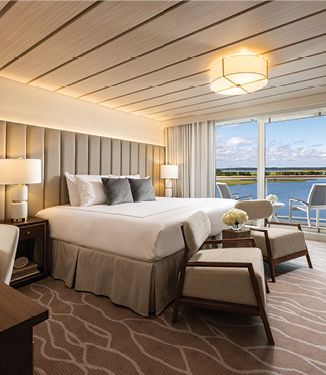 Stateroom with a large bed, comfy chairs, and floor to ceiling windows leading out a balcony.
