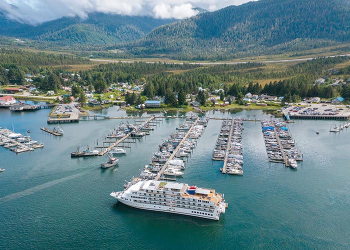 An aerial view of the town of Ketchikan, with several boats docked on the water and the green mountains of Tongass National Forest in the background. 