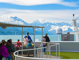 People on a cruise ship deck enjoy scenic views of snow-capped Alaska mountains and a calm blue river.