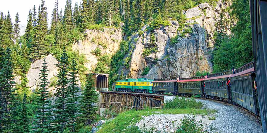 Train on a wooden bridge in a forested, rocky landscape, part of an American Cruise Lines excursion.