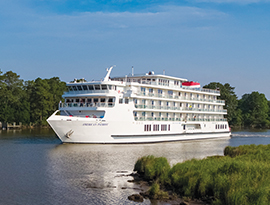The American Patriot, a small American Cruise Lines ship sails on calm Lake Erie, surrounded by lush greenery under a clear blue sky.