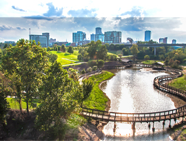 The Arkansas River with a boardwalk in Little Rock, surrounded by lush greenery with the city skyline in the background.