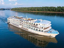 A small American Cruise Lines ship sails on the Arkansas River, surrounded by lush green trees.