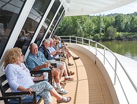 Passengers relax on the deck of an American Cruise Lines ship, enjoying views of lush green riverbanks.