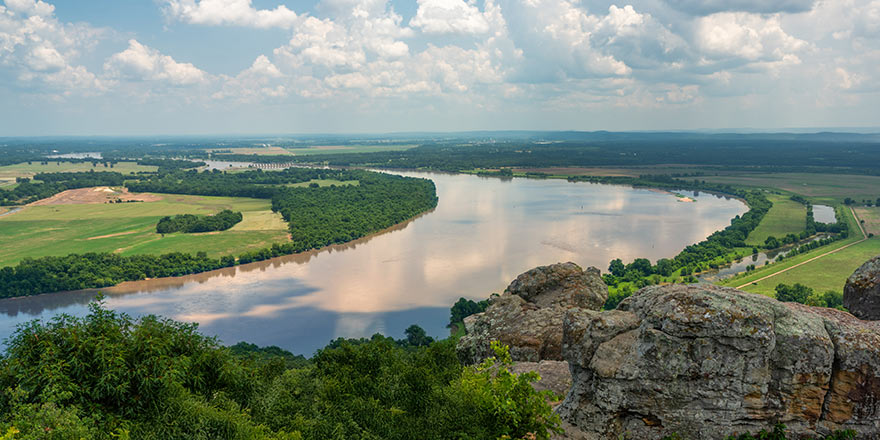 Arkansas River view with lush greenery and rocky foreground, reflecting clouds. Ideal for small ship river cruises by American Cruise Lines.