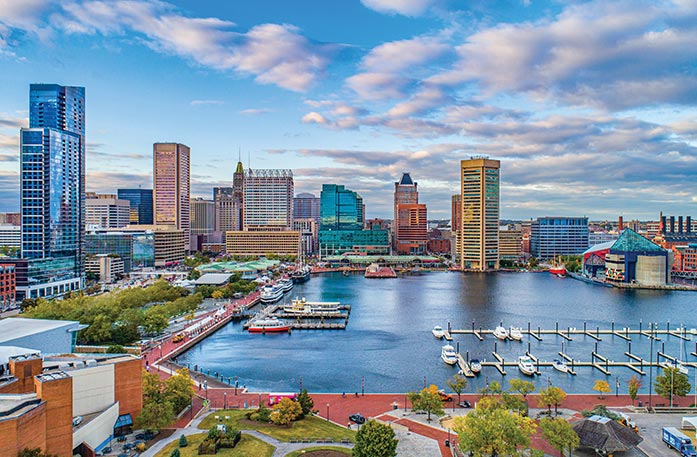 Aerial view of the Baltimore harbor with small cruise ships docked, surrounded by a city skyline with tall buildings and a partly cloudy sky.