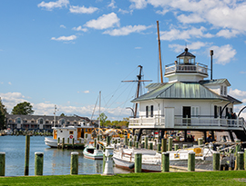 The Choptank River Lighthouse in Cambridge stands on stilts over the water at a marina filled with moored boats along the American Cruise Lines Chesapeake Bay route, featuring a red roof over the white balcony and a small white-and-red boat beneath the balcony.