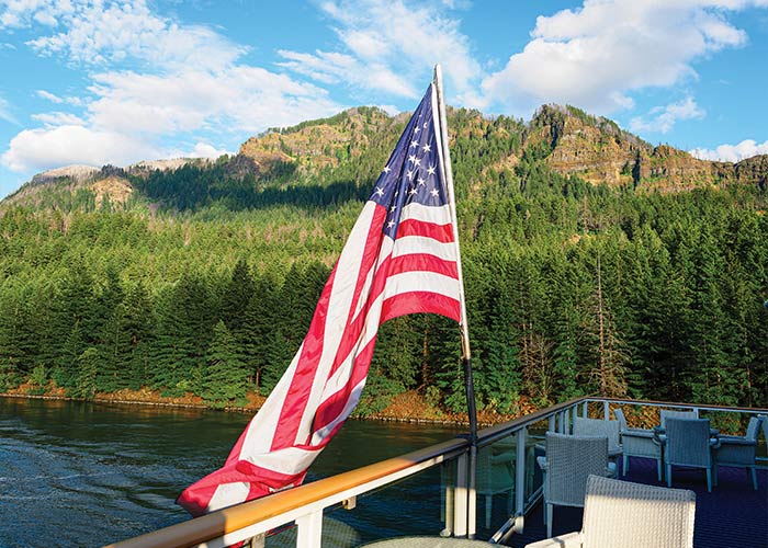 American Song docked along the Columbia River in Richland, where small white flowers peak out from the tall grass. There are trees along the shoreline, a park bench, and mountains in across the river in the distance. In the top left corner, there is a text overlay on a blue box that reads "Brand New Itinerary".