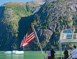 Small Alaska cruise ship with American flag, passengers taking photos of scenic mountains and iceberg.