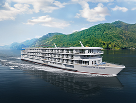 A small American Cruise Lines ship sails on the Columbia River, surrounded by lush green hills under a blue sky.