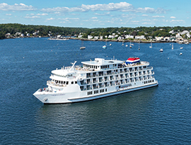 American Patriot cruise ship on one of the Great Lakes, surrounded by scenic shoreline with trees and houses, under a clear blue sky.