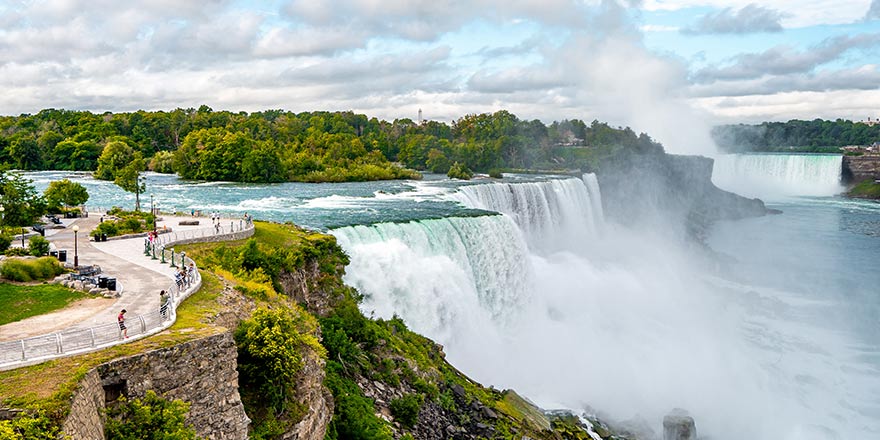 Niagara Falls, a Great Lakes cruise stop, with lush greenery, tourists on a viewing path, and mist rising from the cascading water.