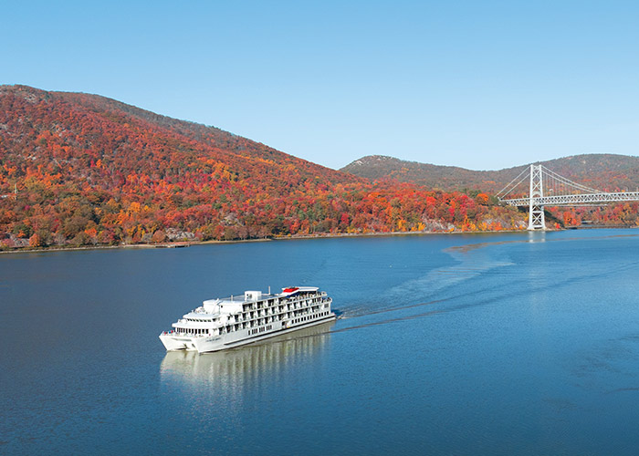 An aerial view of a long bridge across the Hudson River, and the hills covered in trees with vibrant red, orange, and yellow leaves on each side of the river during prime fall foliage season.