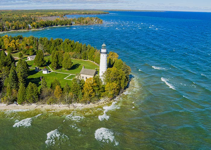 Aerial view of a lighthouse on Michigan's Upper Peninsula, surrounded by blue water, with small waves and distant shoreline, a scenic destination for American Cruise Lines' small ship river cruises.