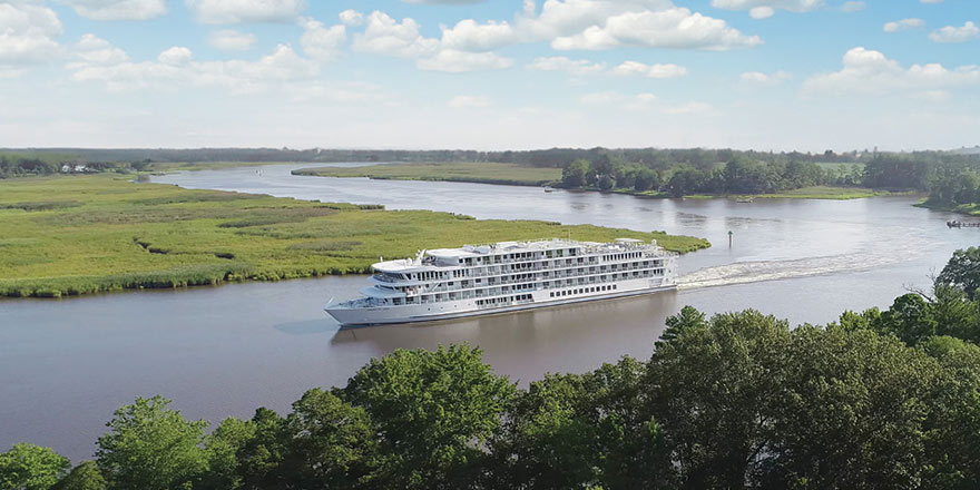An American riverboat gliding along the Mississippi River, surrounded by green landscapes, under a blue sky with a few clouds.