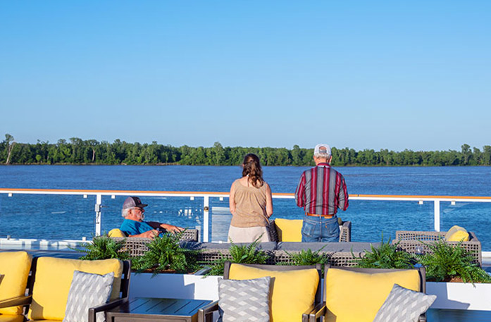 On the deck of the American Heritage paddlewheeler, there is a large American flag, with it's pole on the glass railing, blowing in the wind with the views of the Mississippi River. The floor of the deck is dark blue, a few stairs can be seen to the left, and there is an empty tan colored chair and table in the foreground.