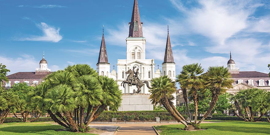 The St. Louis Cathedral with three dark grey spires stands in Jackson Square in New Orleans, surrounded by bright green trees and bushes.