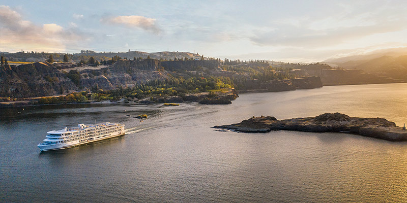 An aerial view of an American Riverboat cruising through the Columbia River Gorge, where there is a small island formation in the middle of the water. Alongside the water, there are several cliffs, and mountains in the distance.