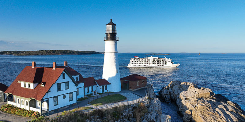Portland Head Light, a white lighthouse on top of a cliff alongside Casco Bay in Portland with a large white building with a dark red roof to the left of the lighthouse. An American Cruise Lines small ship is sailing by in the background.
