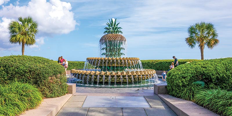 The Pineapple Fountain, a large fountain with water flowing down from layers that resemble a pineapple split in three sections, with the crown as the top layer, in Charleston Waterfront Park. Perfectly trimmed bushes surround the circular pool below the fountain, and there are two palm trees on each side.