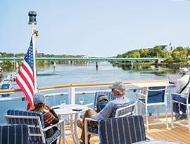 Passengers relax on an American Cruise Lines ship deck with an American flag, overlooking a scenic river and bridge, surrounded by lush greenery.