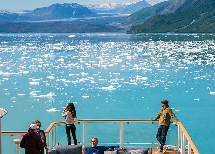 A large field of bright pink Fireweed wildflowers leads up to a thick forest. The snow-capped mountains of Mendenhall Glacier are in the distance.
