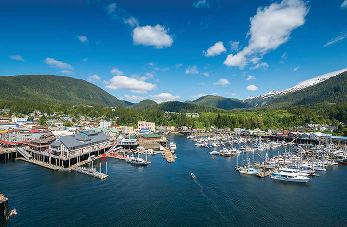 Scenic harbor in Ketchikan, AK with small cruise ships, sailboats, and a coastal town surrounded by lush green mountains.