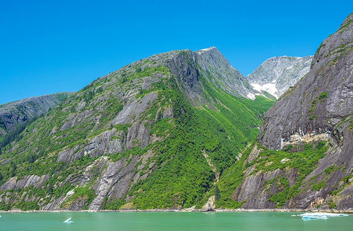 The American Constellation Coastal Cruise Ship sailing through the light green colored passage of Tracy Arm Fjord, surrounded by a cliff with a narrow waterfall.