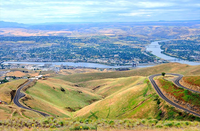 An aerial view of the city of Lewiston. In the foreground, there are grassy hills that lead up to the large cityscape in the center. In the far distance, there are several mountains spanning several miles. 