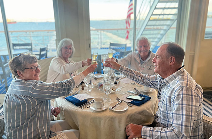 A couple sits at a table beside an American flag on an American Riverboat's sun deck looking out at the Mississippi River, while a man sits to the right of them typing on his computer.