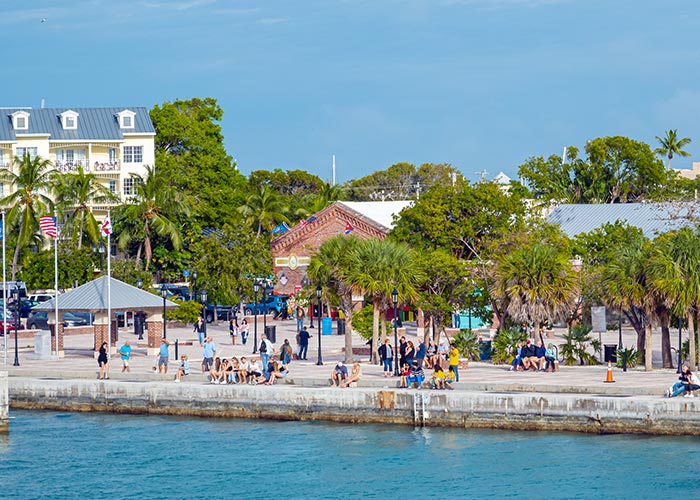 A bottom-up view of Key West Lighthouse & Keeper's Quarters, a white lighthouse with oak trees standing beside it under a blue sky at the Key West Art & History Museum.