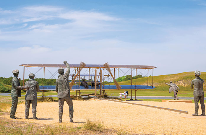 A recreation of the first flight, with a large sculpture of an airplane with a statue of a person laying on it, and four statues on the ground behind it at Wright Brothers National Memorial in Kitty Hawk.
