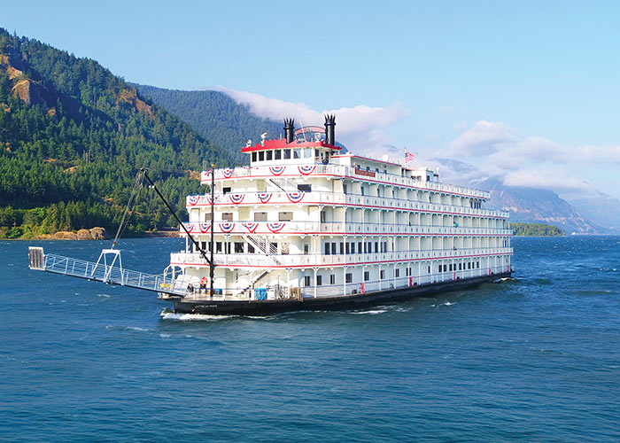 American Pride, an American Cruise Lines Paddlewheeler sailing along the Columbia River. Alongside the river, there are several cliffs covered in thick greenery, and silhouettes of mountains in the distance.