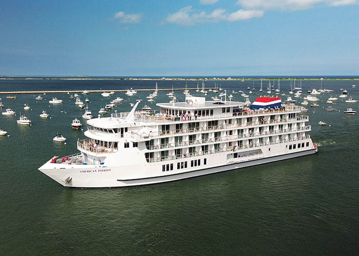 An aerial view of American Patriot, an American Cruise Lines Patriot Class small cruise ship sailing along the New England coast, with views of the shore in the distance.