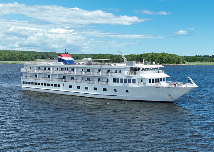 American Independence, an American Cruise Lines small ship departing from a small New England harbor, where there are several small boats docked along the water. Trees and buildings cover the shoreline, and small mountains fill the horizon.