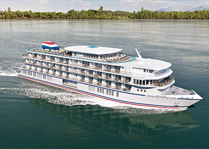 An aerial view of American Maverick, an American Cruise Lines Patriot Class small cruise ship sailing along the New England coast, with views of the shore in the distance.