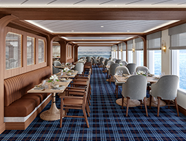 The restaurant on American Anthem, where there are several round tables and navy blue chairs along the blue and white patterned carpet, and two round chandeliers hanging from the ceiling. There are large windows with clear views of the Columbia River on one wall, and floor-to-ceiling paintings on the back wall.