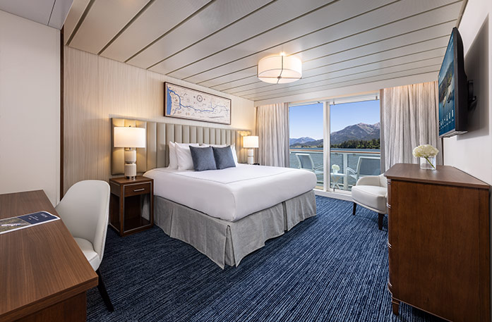 The Vista Balcony stateroom on American Encore, with large floor-to-ceiling sliding glass doors, with curtains on each end that lead to a private balcony with a table and chairs overlooking the Columbia River. There is an end table with a lamp on each side of the bed. 