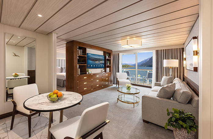 The Grand Suite on American Encore, with large floor-to-ceiling sliding glass doors leading to a private balcony overlooking the Columbia River. There is a grey sofa and an armchair facing a large wooden TV stand in the center of the room, with a TV and shelves with decor. 